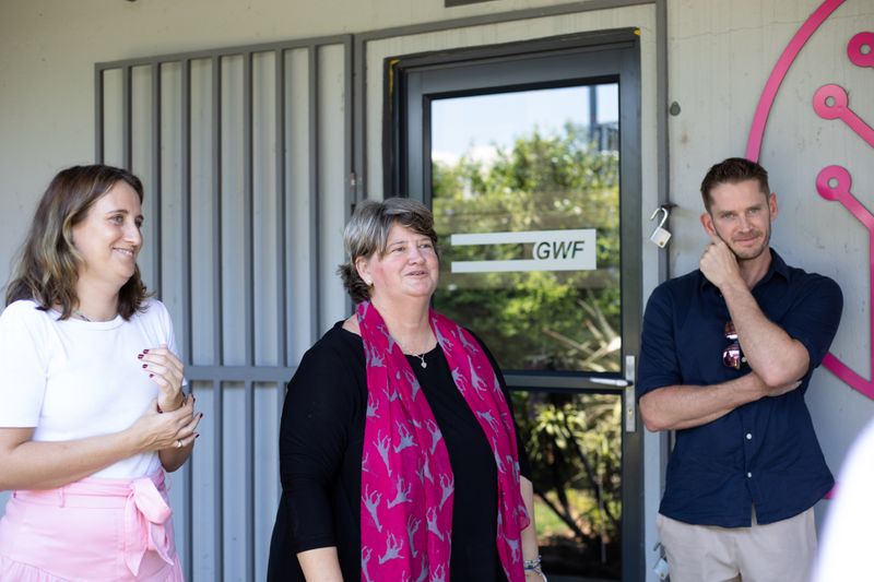 Three individuals stand outside a GWF building, listening attentively, with one woman in a black outfit and a pink scarf speaking passionately while the others react with smiles and thoughtfulness.