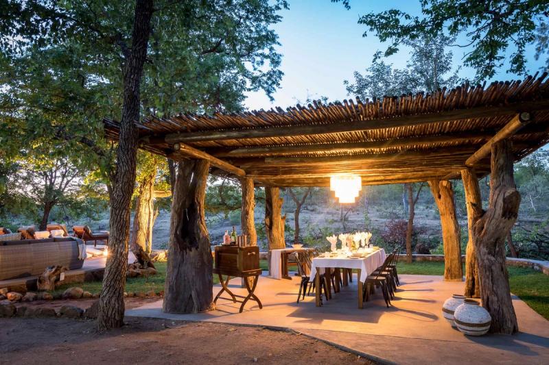 Comedor al aire libre bajo una pérgola de madera, rodeado de árboles y bañado por la luz cálida del atardecer.