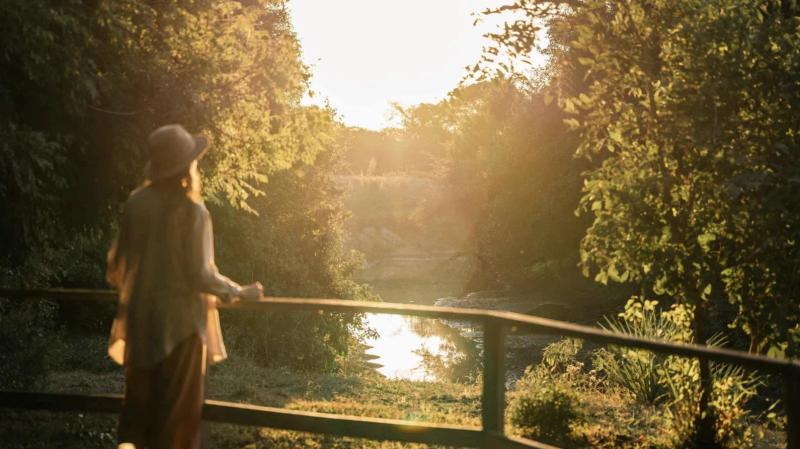 Person standing on a wooden walkway overlooking the river and forest at sunset.