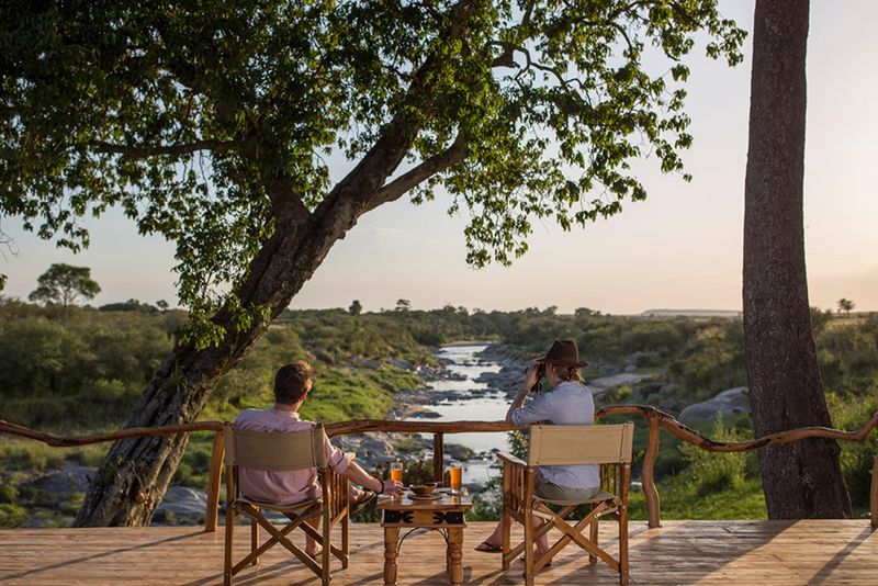 Two people relax on a wooden deck overlooking a serene river surrounded by lush greenery, with drinks on a small table between them