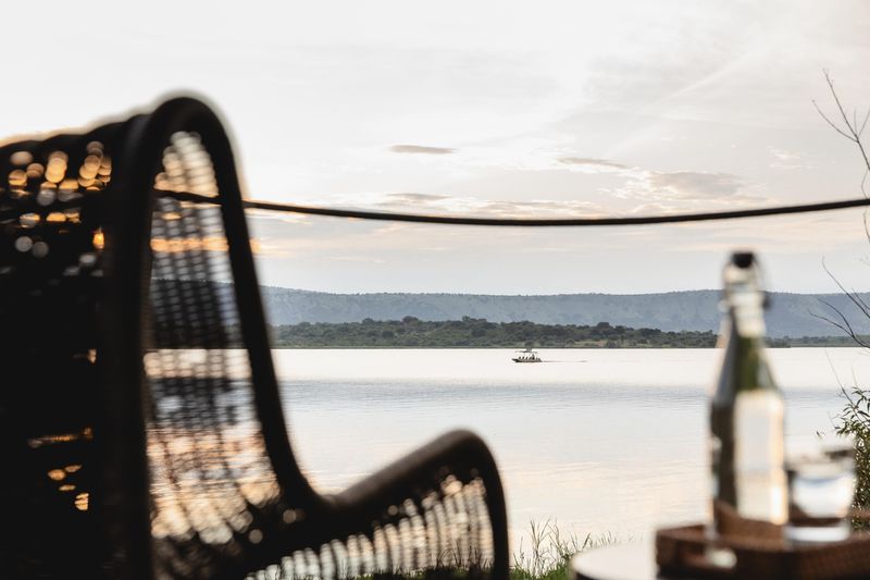 A boat glides across a still lake at sunset, viewed through the frame of a wicker chair and a table with a bottle and glass.