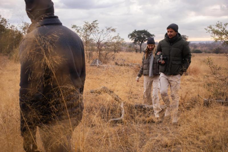 A couple dressed in warm outdoor clothing walks arm-in-arm through dry grassland, following a guide during a guided nature walk in an African bush landscape under a cloudy sky.