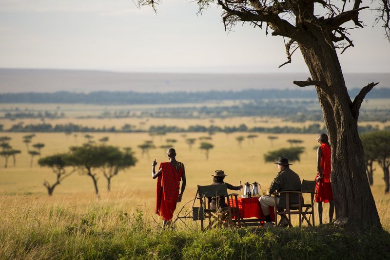 A small group enjoys an outdoor meal under a tree, with Maasai guides nearby and vast savannah stretching into the distance