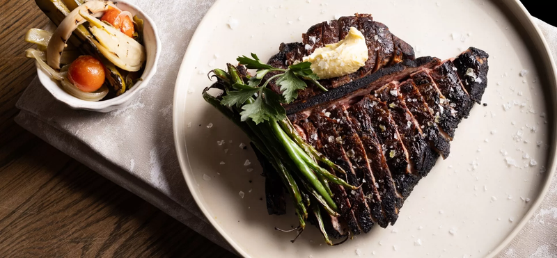Top-down view of a sliced grilled bone-in steak topped with melting butter and served with charred green beans on a white plate, beside a small bowl of roasted vegetables.