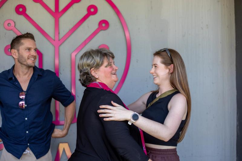 A joyful moment is captured as two women warmly embrace, their smiles radiating genuine connection, while a man in the background watches with an amused expression.