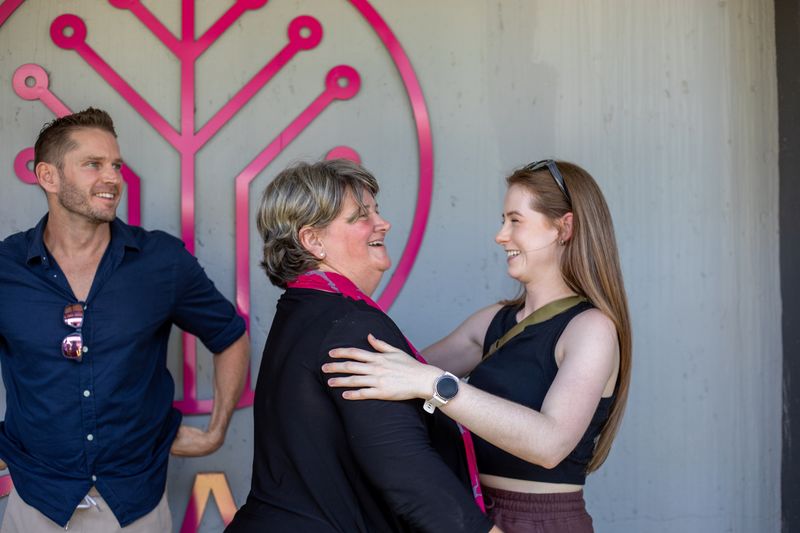 A joyful moment is captured as two women warmly embrace, their smiles radiating genuine connection, while a man in the background watches with an amused expression.