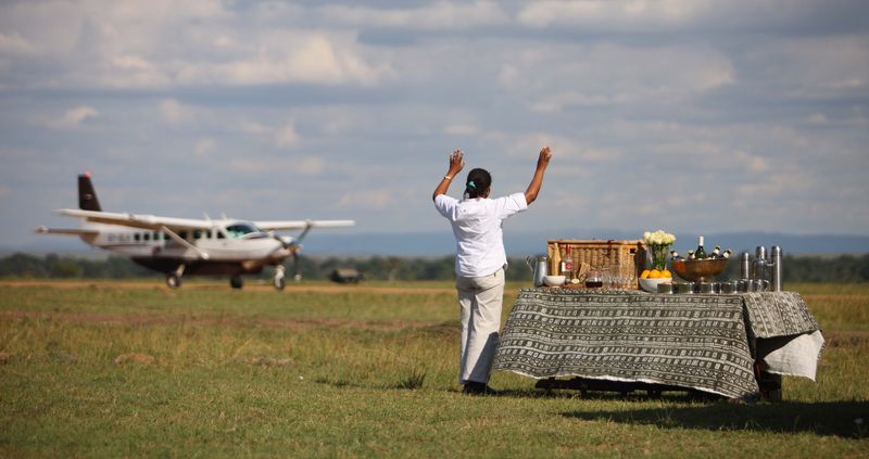 A camp staff member waves to arriving guests stepping off a small bush plane beside a table laid out with refreshments on a grassy airstrip.