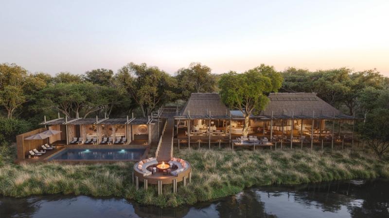 An elevated riverside lodge with open-sided decks, a pool, and a circular fire pit sits nestled among trees overlooking the Khwai River.