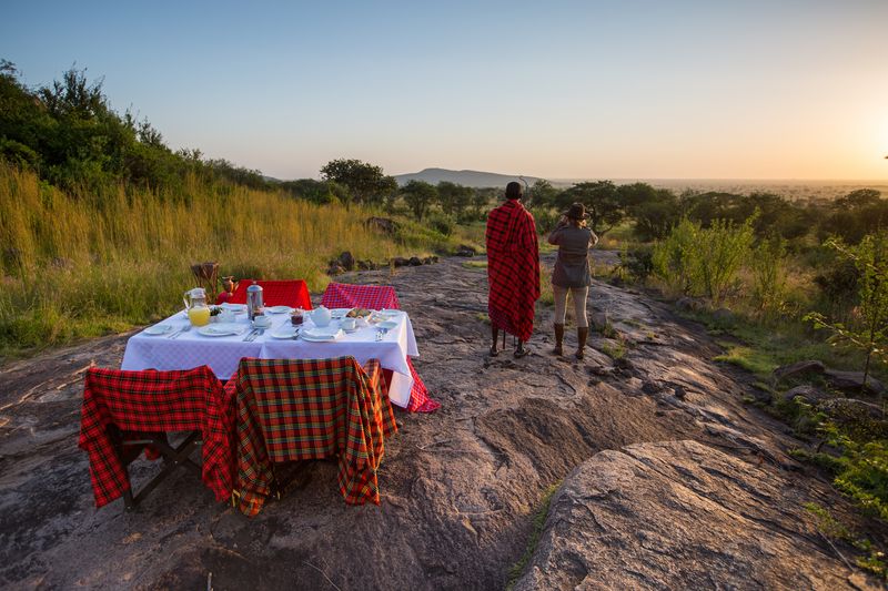 Guests enjoy a Serengeti Pioneer Camp bush breakfast on a rocky outcrop, overlooking the plains at sunrise with a Maasai guide nearby.