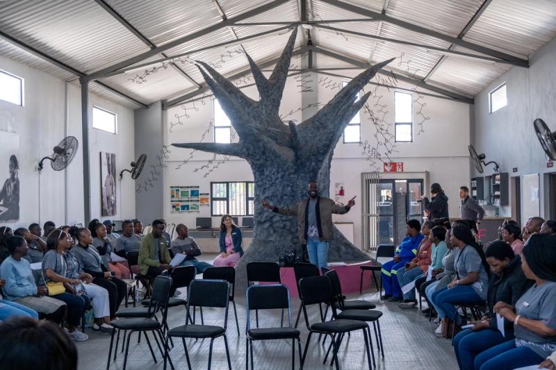 A speaker stands confidently in front of a group of engaged listeners, gesturing animatedly, with a striking tree sculpture behind him as a symbol of growth and learning.