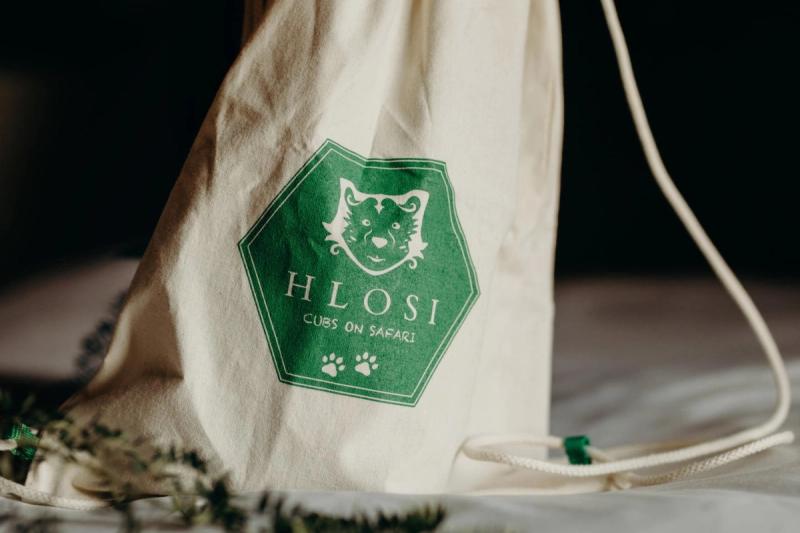 A drawstring bag with the green logo of "Hlosi Cubs on Safari," featuring a lion's face and paw prints, is placed on a soft surface with subtle foliage in the foreground.