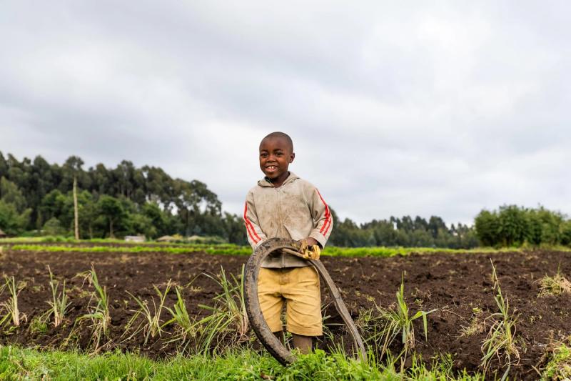 A child smiles standing in front of a farm in Rwanda