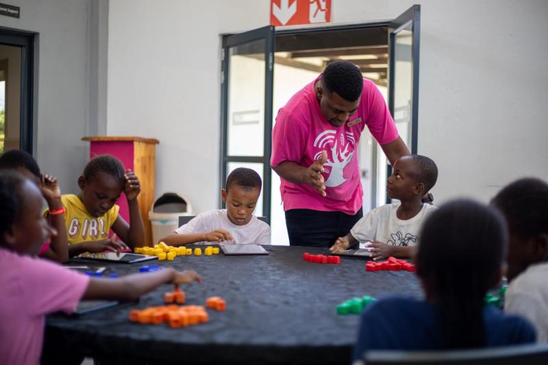 A passionate teacher in a bright pink shirt leans in to engage with a young student, as children around a table work with tablets and colourful counting blocks in a lively learning environment.
