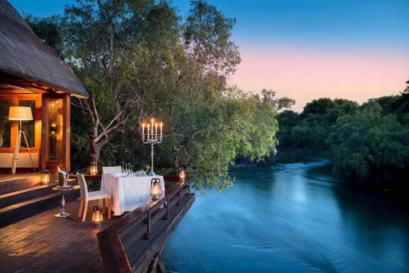 A romantic dining setup on a wooden deck overlooking a serene river, with a candelabra centerpiece, lanterns, and soft lighting against a backdrop of lush greenery and a twilight