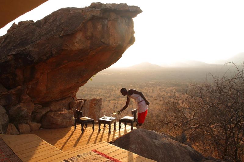 A Maasai man setting up drinks on a small wooden table under the shade of a large rock outcrop, with a vast, sunlit savannah stretching into the horizon