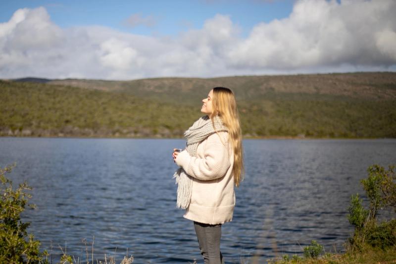 Katharina stands by a tranquil lake, wrapped in a warm scarf, eyes closed as she tilts her face toward the sun during her babymoon safari.