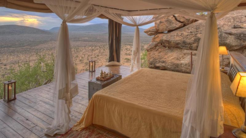 An elegant open-air bedroom with a four-poster bed draped in soft white netting, set on a wooden deck with lanterns, overlooking a vast savannah landscape surrounded by rocky outcrops