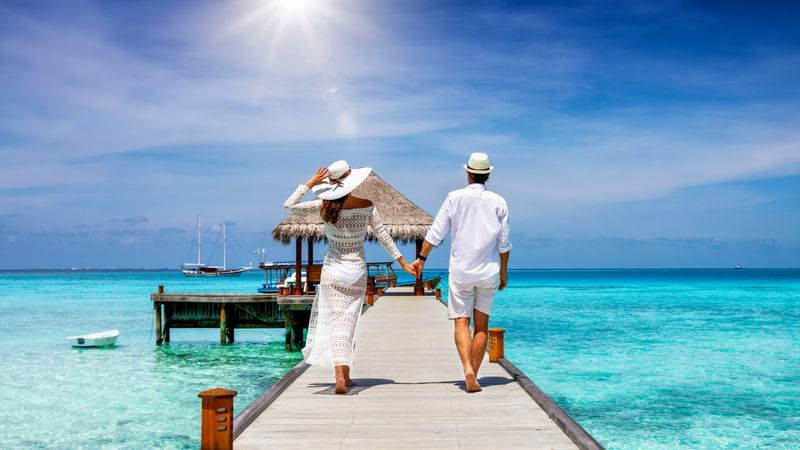 A couple dressed in white walks hand-in-hand along a wooden pier over turquoise water, heading toward an overwater bungalow under a sunny sky