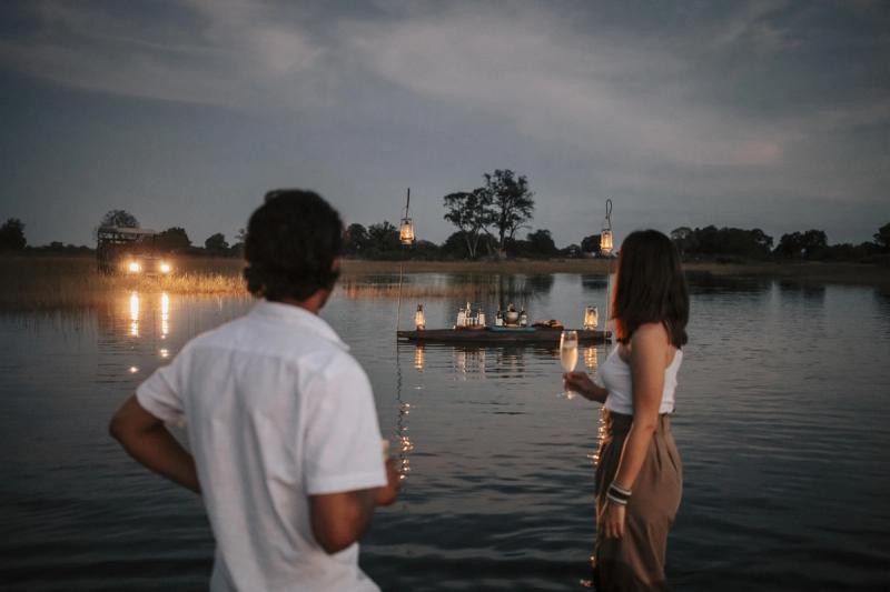 A couple stands ankle-deep in water, champagne in hand, gazing at a floating lantern-lit bar in the Okavango Delta—an unforgettable way to enjoy the best time to visit Botswana for a safari.