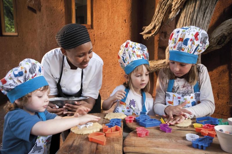 A group of young children wearing colourful chef hats and aprons excitedly learn baking skills from a smiling chef, working together at a wooden table outdoors.