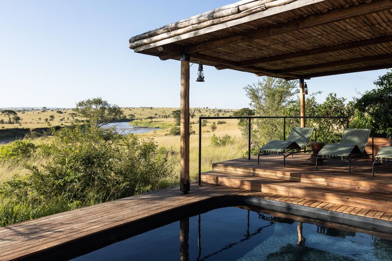 A private wooden deck with loungers and a plunge pool overlooks a winding river and golden plains, capturing the seamless indoor-outdoor design typical of luxury safari lodges in Tanzania.