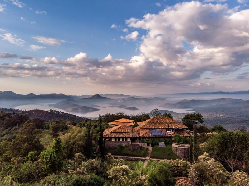 A stunning lodge with terracotta roofs sits atop a lush hillside, overlooking misty lakes and rolling mountains under a dramatic cloudy sky in Rwanda
