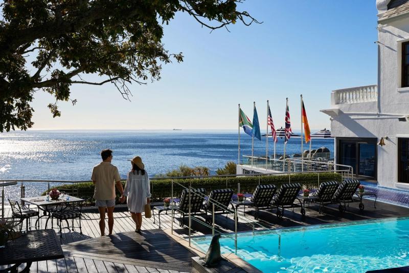 A couple on on a romantic safari in Africa walking hand-in-hand by a pool deck adorned with sun loungers, overlooking the sparkling Atlantic Ocean, with flags from various countries fluttering in the breeze and a bright blue sky above
