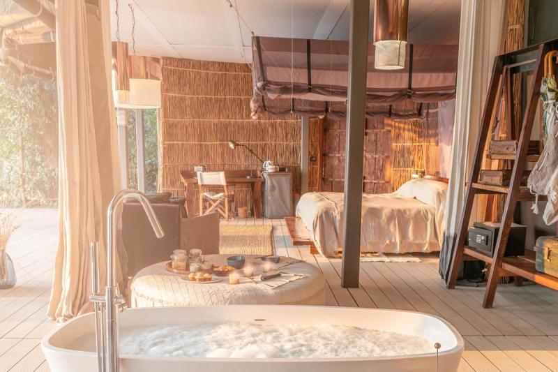 A luxurious safari-style suite with a freestanding bubble-filled bathtub, a cosy bed under a canopy, rustic furnishings, and warm natural tones illuminated by soft natural light