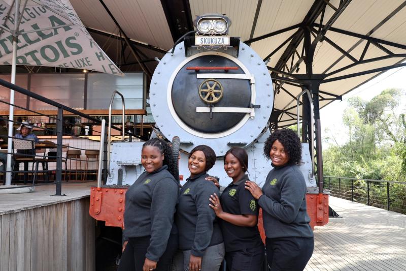 Image of Lwazi Thobela, Noxolo Mathebula, Vutivi Mthimkhulu and Mampho Makofane posing in front of a stationary train at the Kruger Station restaurant