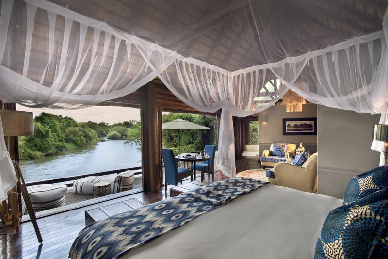 A luxurious riverside suite with a canopy-draped bed, elegant seating, and an open deck overlooking a tranquil river surrounded by lush greenery.