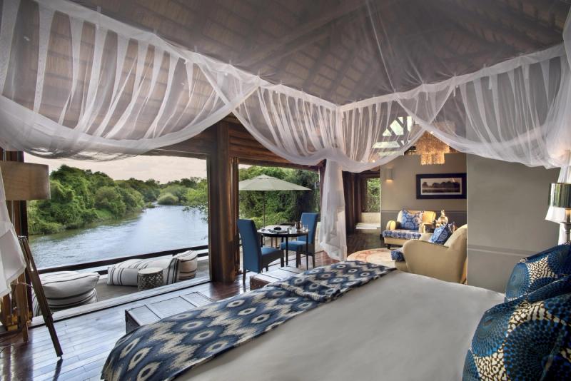 A luxurious riverside suite with a canopy-draped bed, elegant seating, and an open deck overlooking a tranquil river surrounded by lush greenery.