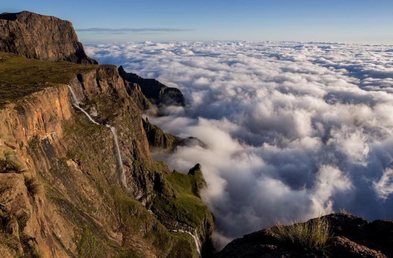 Mist rolls through the dramatic cliffs of the Drakensberg, with lush green slopes leading down to a vast, open valley.