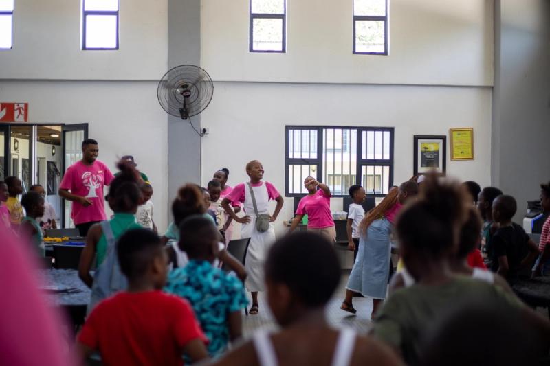 A room filled with children and educators brims with energy as facilitators in pink shirts lead an animated activity, their enthusiasm reflected in the excited faces of the students.