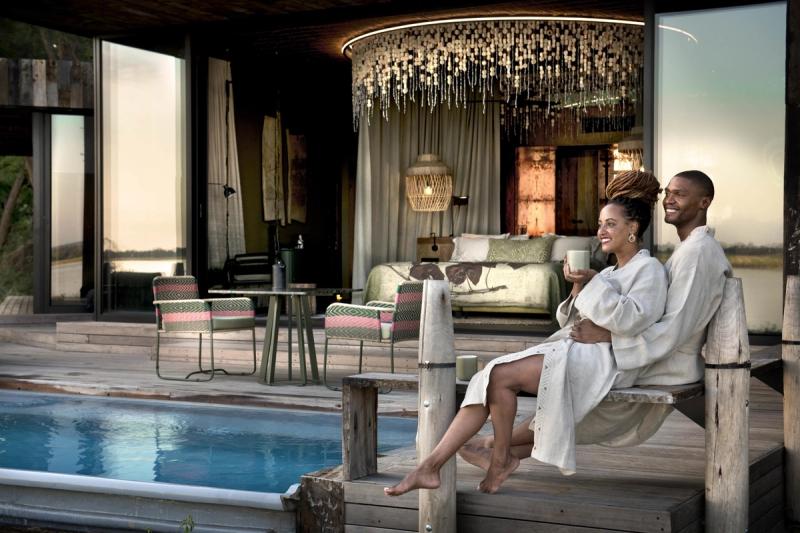 A joyful couple in robes relaxing by a private pool at a luxurious, open-plan safari lodge with elegant decor and tranquil surroundings