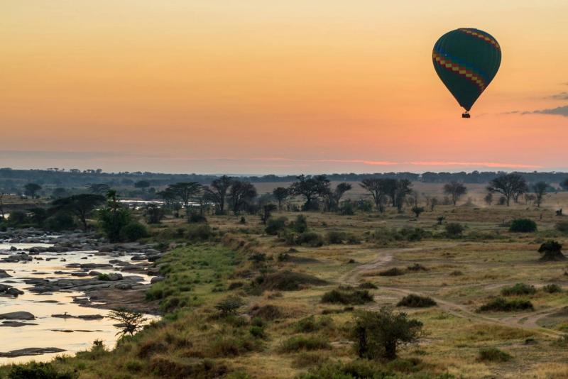 A hot air balloon safari over the Serengeti's vast plains at sunrise, with a rocky river and scattered acacia trees in the foreground