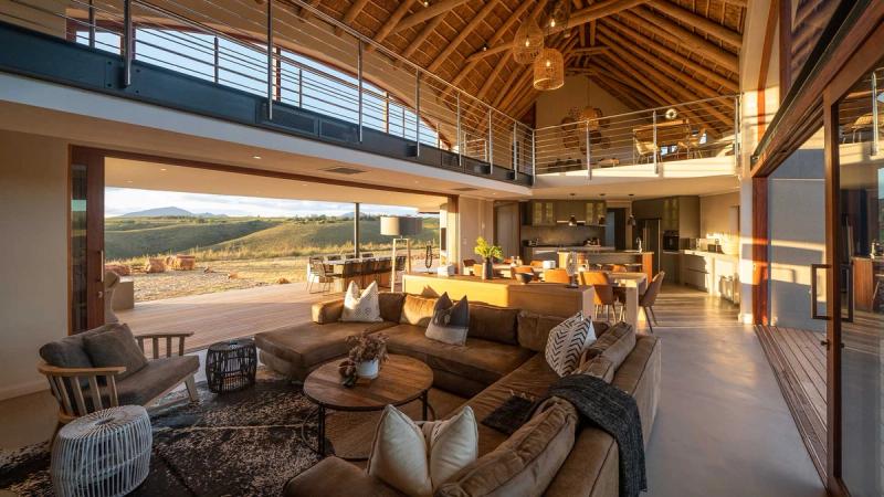 A spacious, open-plan lodge interior with modern furnishings, a high thatched ceiling, and large glass doors offering a view of rolling hills bathed in golden sunlight.