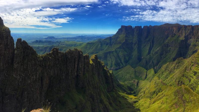 The sun highlights the towering cliffs of the Drakensberg Mountains, casting light over the rugged peaks and lush valleys below.