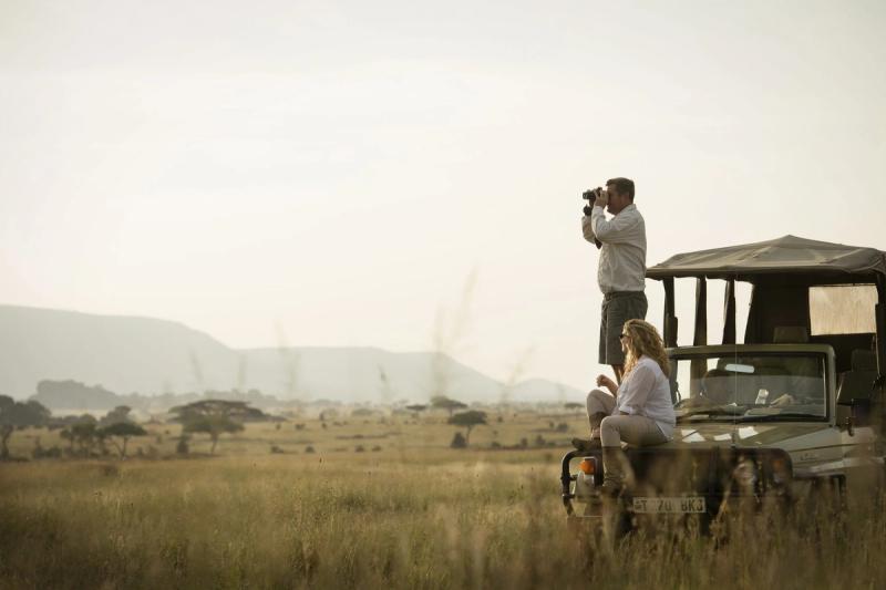 Guests on a Dunia Camp game drive vehicle in Tanzania’s Serengeti, with one person standing to photograph the open savannah while another sits on the bonnet overlooking the plains.
