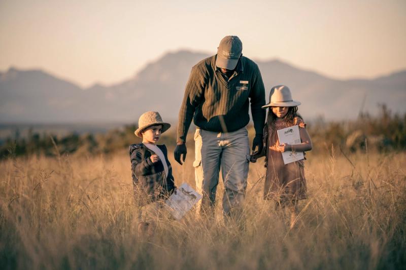 A ranger guides two young children through a grassy field at sunset, with mountains in the background. The children hold safari activity sheets and wear wide-brimmed hats.