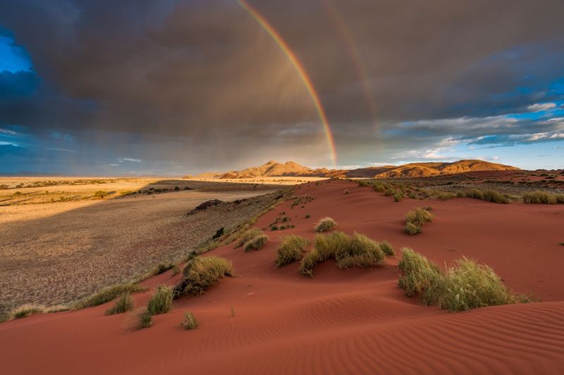 A double rainbow arcs over Namibia’s red dunes and distant mountains, capturing a manifesto moment shaped by light and wide-open silence.