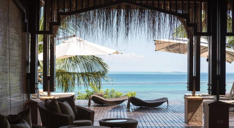 A shaded seating area with wicker chairs and cushions, opening onto a wooden deck with modern loungers and white umbrellas, all overlooking a turquoise ocean framed by palm trees.