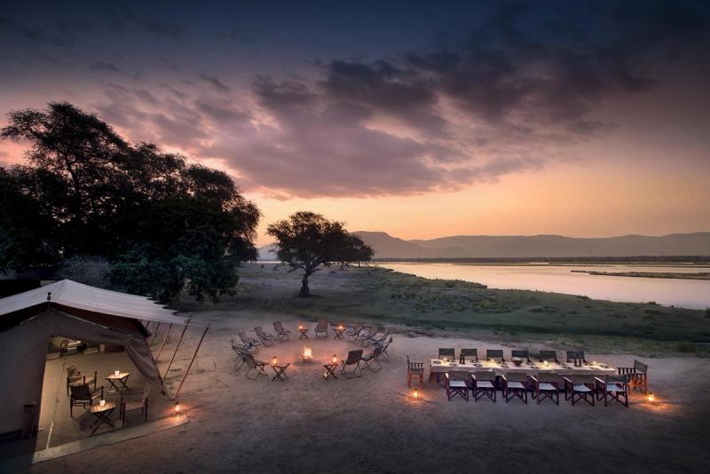 An open-air safari camp beside the Zambezi River at sunset, featuring a campfire circle, a long dining table, and a canvas tent