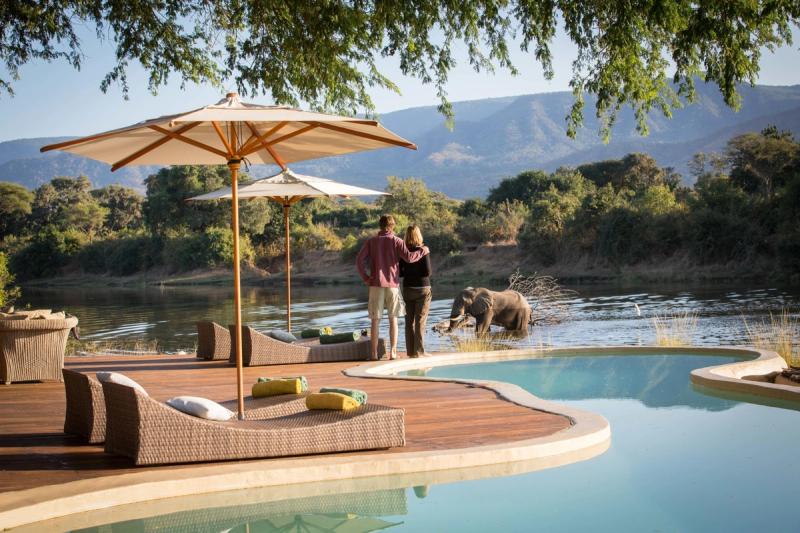 A couple enjoys a peaceful moment by an infinity pool overlooking a river, as an elephant wades nearby amidst a stunning wilderness backdrop