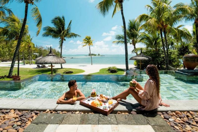 A couple enjoying a floating breakfast in a luxurious pool overlooking a tropical beach lined with palm trees under a bright blue sky