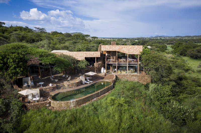 An elevated stone-and-glass safari lodge with a curved infinity pool overlooks vast green plains and scattered acacia trees, capturing the open, contemporary feel of Luxury safari lodges in Tanzania.