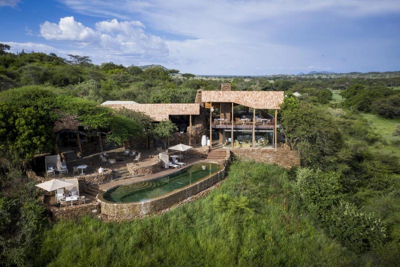 An elevated stone-and-glass safari lodge with a curved infinity pool overlooks vast green plains and scattered acacia trees, capturing the open, contemporary feel of Luxury safari lodges in Tanzania.