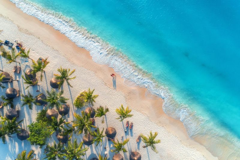 An aerial view of a tropical beach in Zanzibar, showing waves lapping against the shore, rows of umbrellas, and the turquoise sea at sunset.