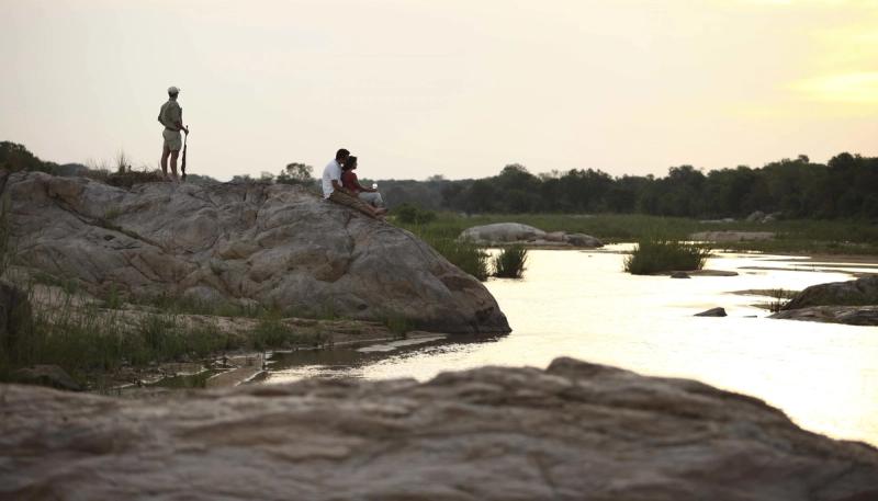 A couple seated on a large rock by a tranquil river, enjoying the serene wilderness, with a guide standing nearby overlooking the peaceful landscape