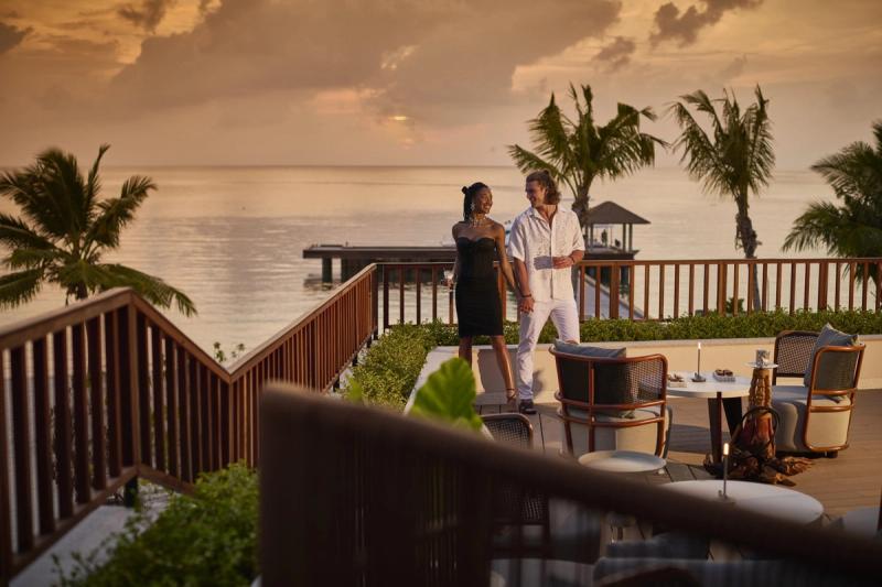 A couple strolling hand in hand on a stylish outdoor terrace overlooking a calm ocean at sunset, framed by palm trees and elegant seating