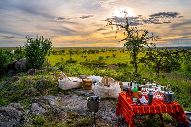 A scenic sundowner setup with beanbags and a table of drinks overlooks the vast green plains of the Serengeti under a golden sunset sky